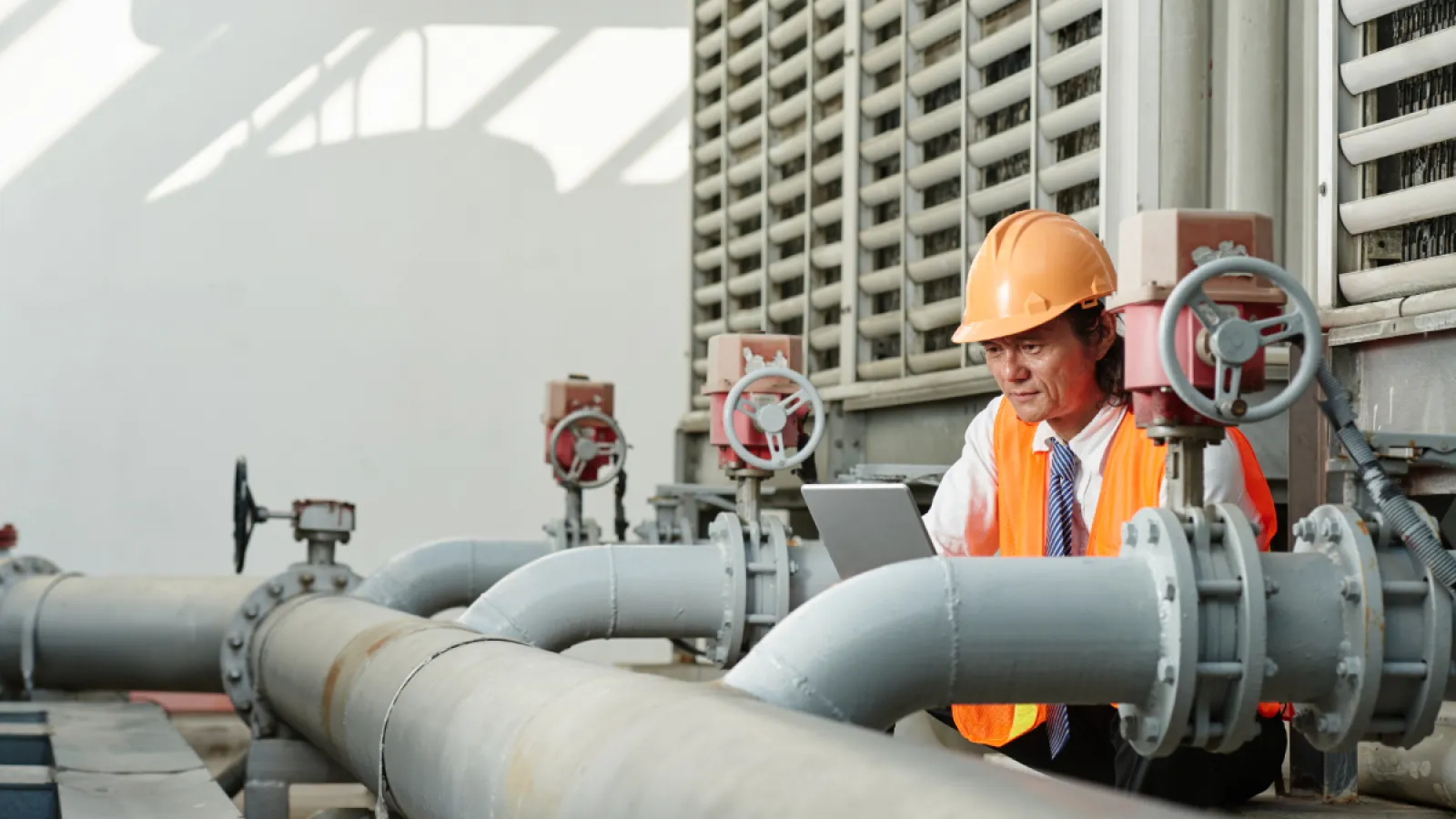 Engineer in safety gear inspecting industrial pipes and valves while using a tablet in a plant facility.