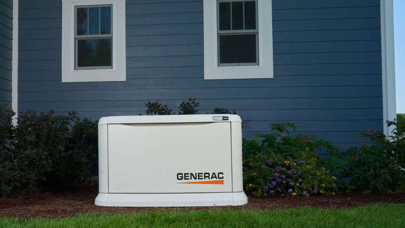 Generac home standby generator installed outside a blue house with green lawn and flowers nearby