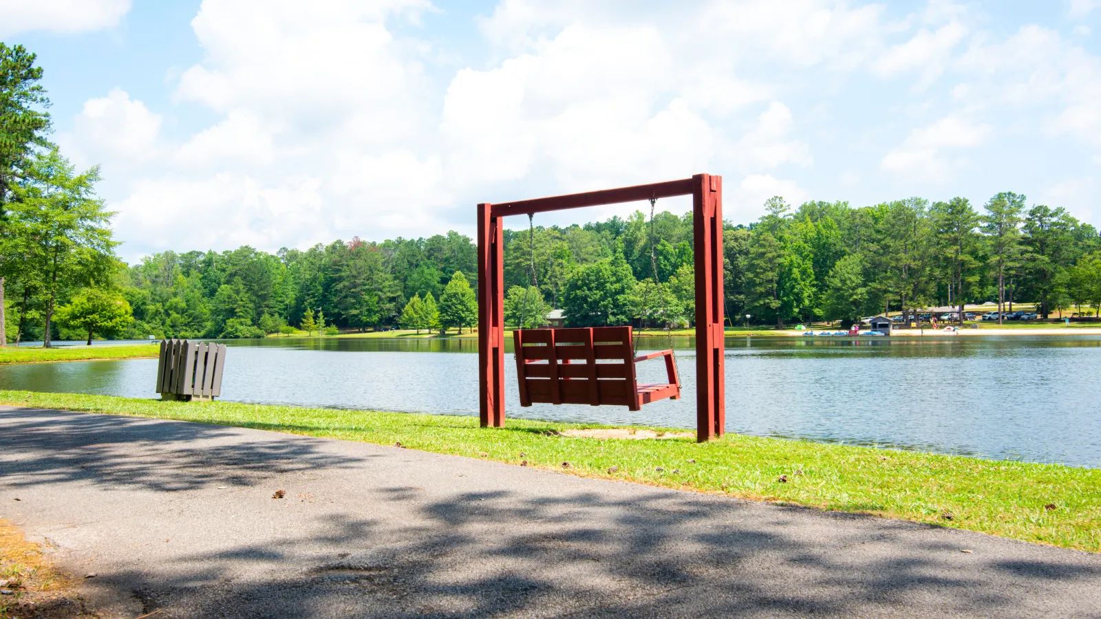 Red wooden swing bench by a lakeside path with green trees and a partly cloudy blue sky.
