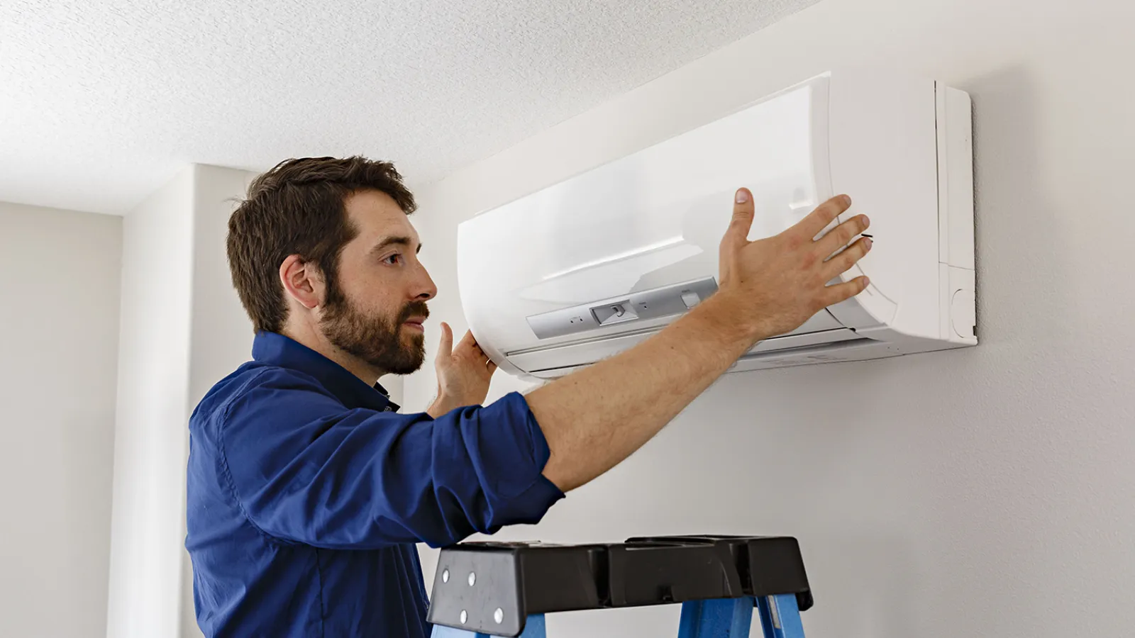 Technician in blue shirt installing a white wall-mounted air conditioner unit using a blue ladder.