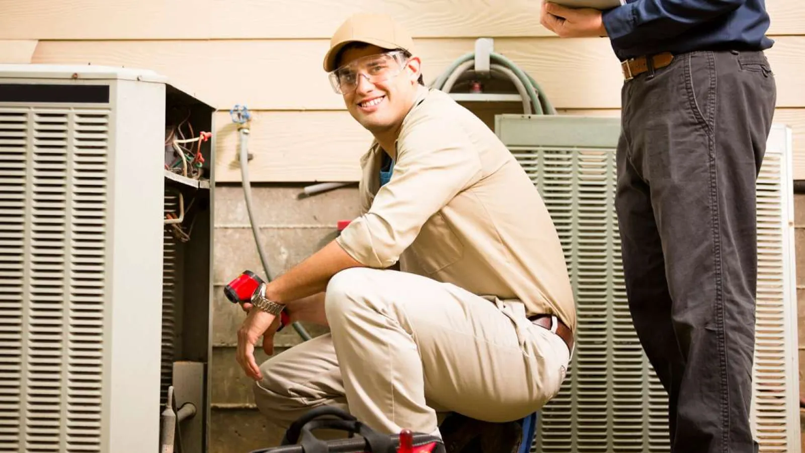 HVAC technician in beige uniform repairing outdoor air conditioner unit while client watches.