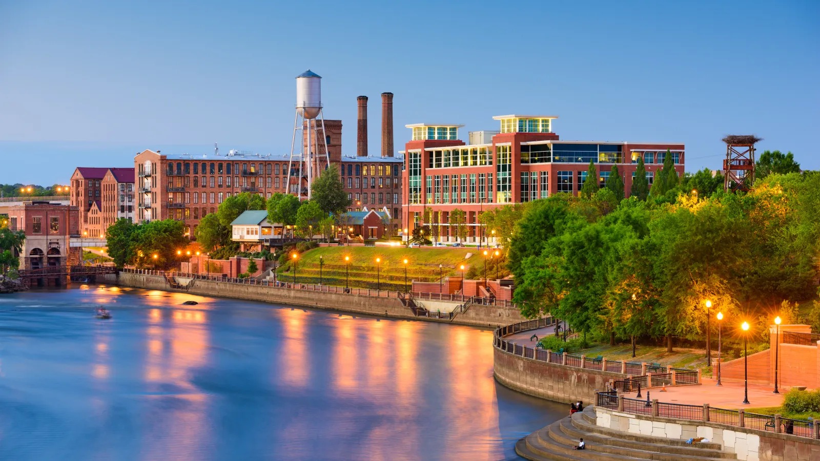 Evening view of historic industrial buildings and water tower along a river with illuminated waterfront park.