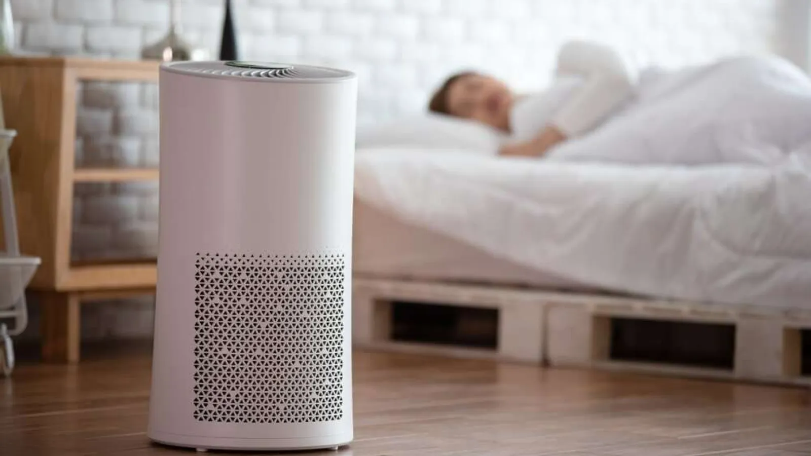 Modern white air purifier on wooden floor with sleeping person in bed in background for clean air concept