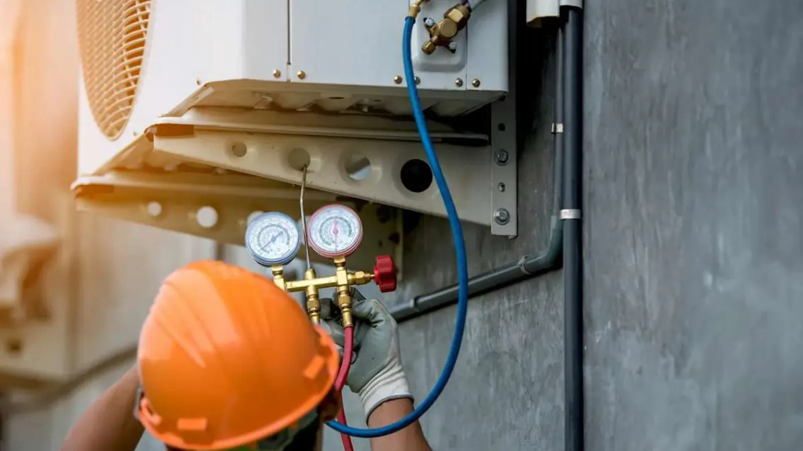 Technician in orange helmet adjusting air conditioner gauges with blue and red hoses on wall-mounted HVAC unit.