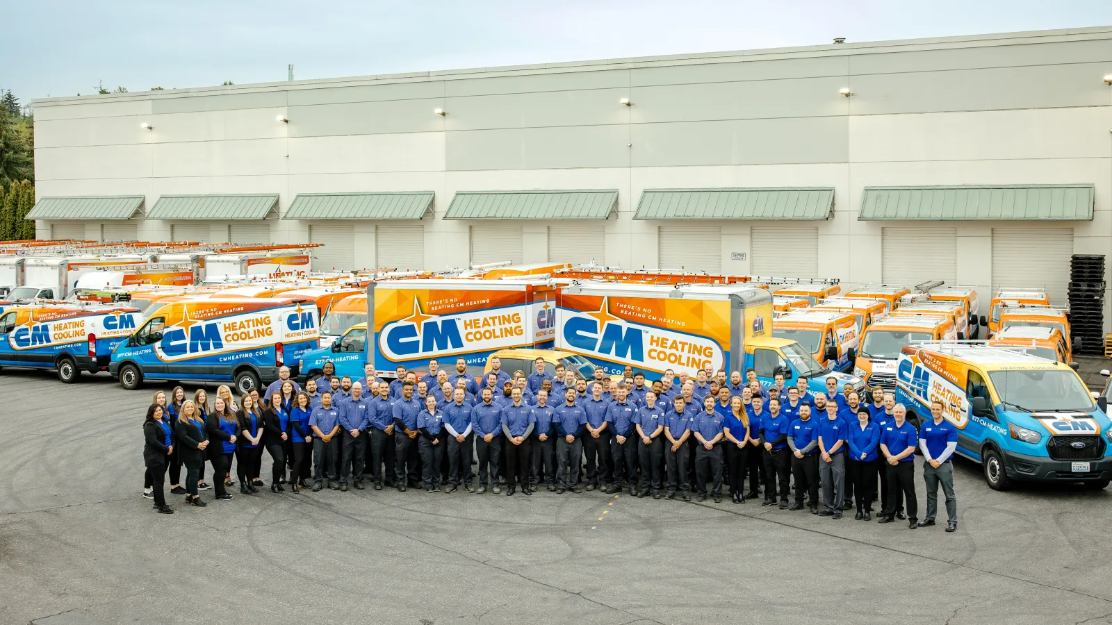 Large group of CM Heating and Cooling employees standing in front of branded service trucks outside warehouse