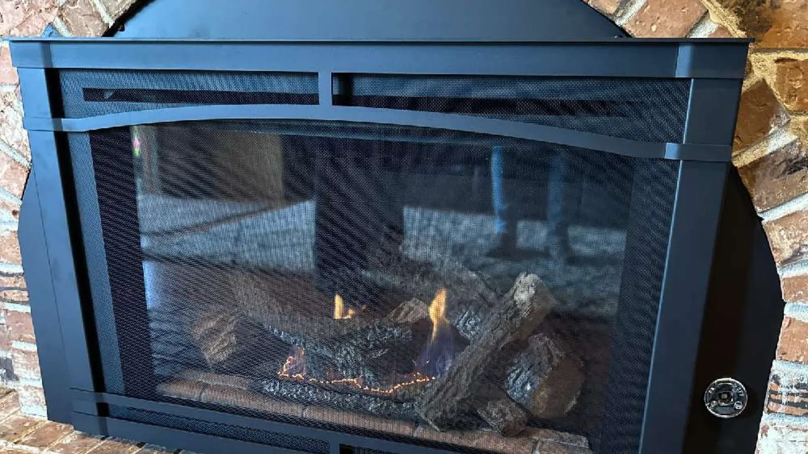 Modern black fireplace with burning logs and protective mesh cover set in brick wall.