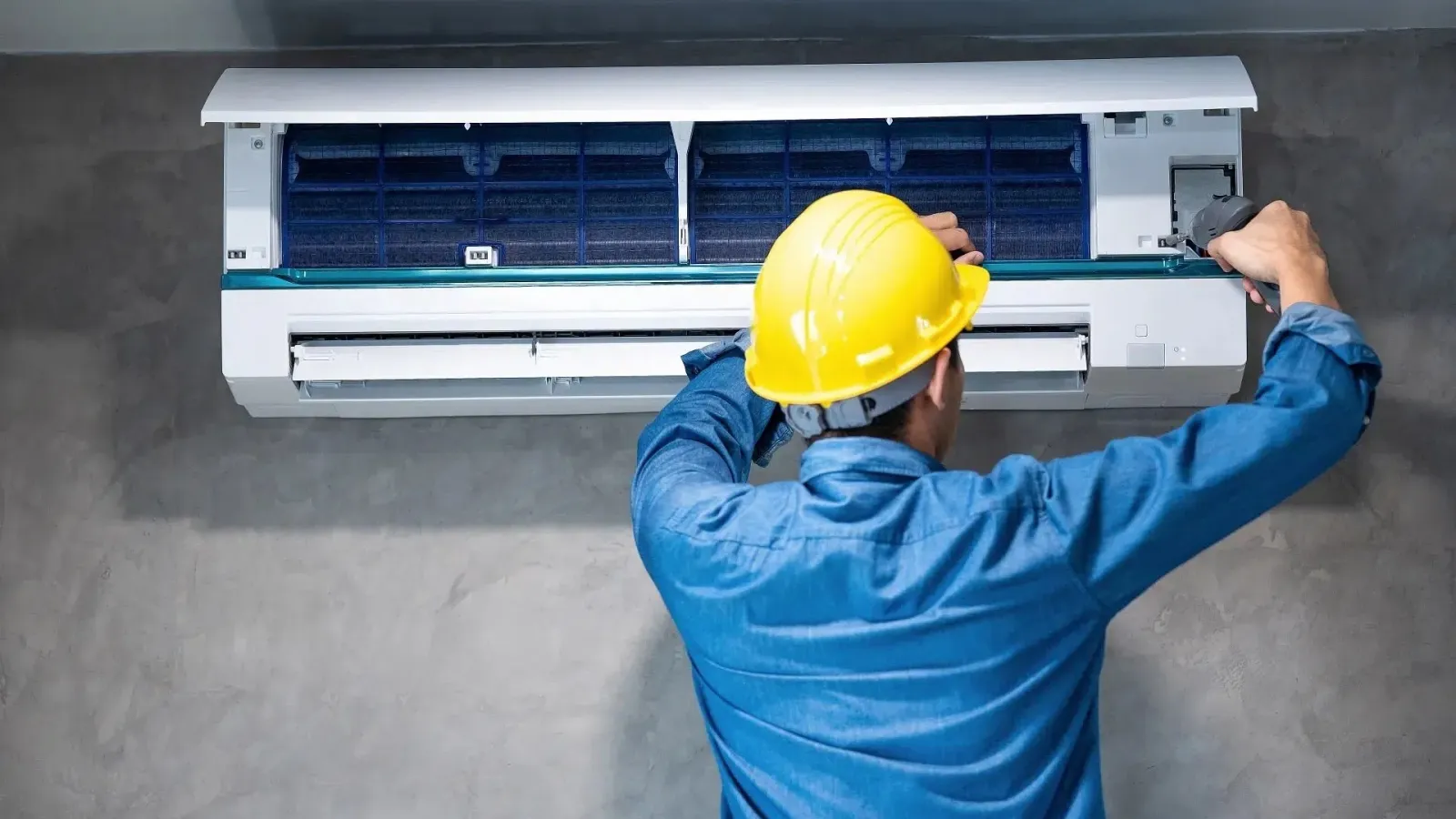 Technician in a yellow helmet repairing a wall-mounted air conditioning unit indoors against a gray wall.