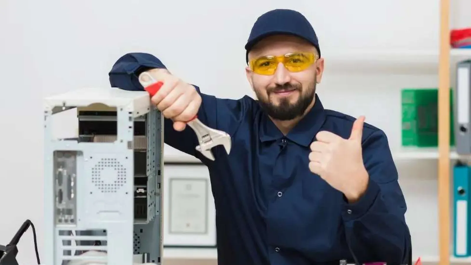 Technician in protective glasses showing thumbs up while repairing computer hardware with a wrench.