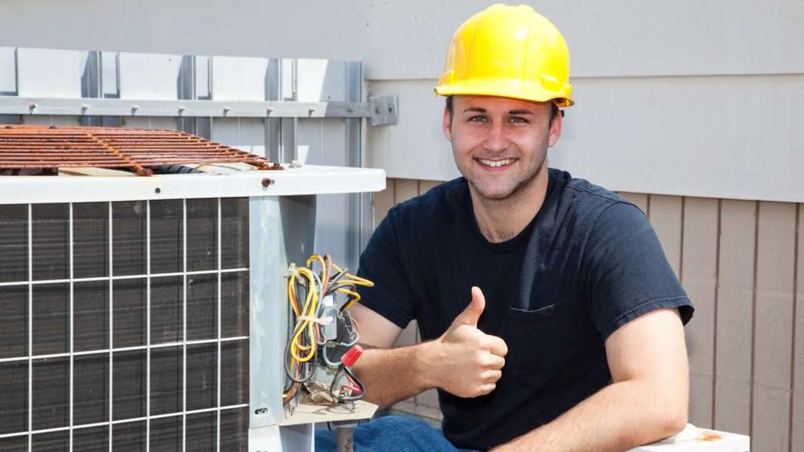 Technician wearing yellow hard hat giving thumbs up next to outdoor air conditioning unit during maintenance.