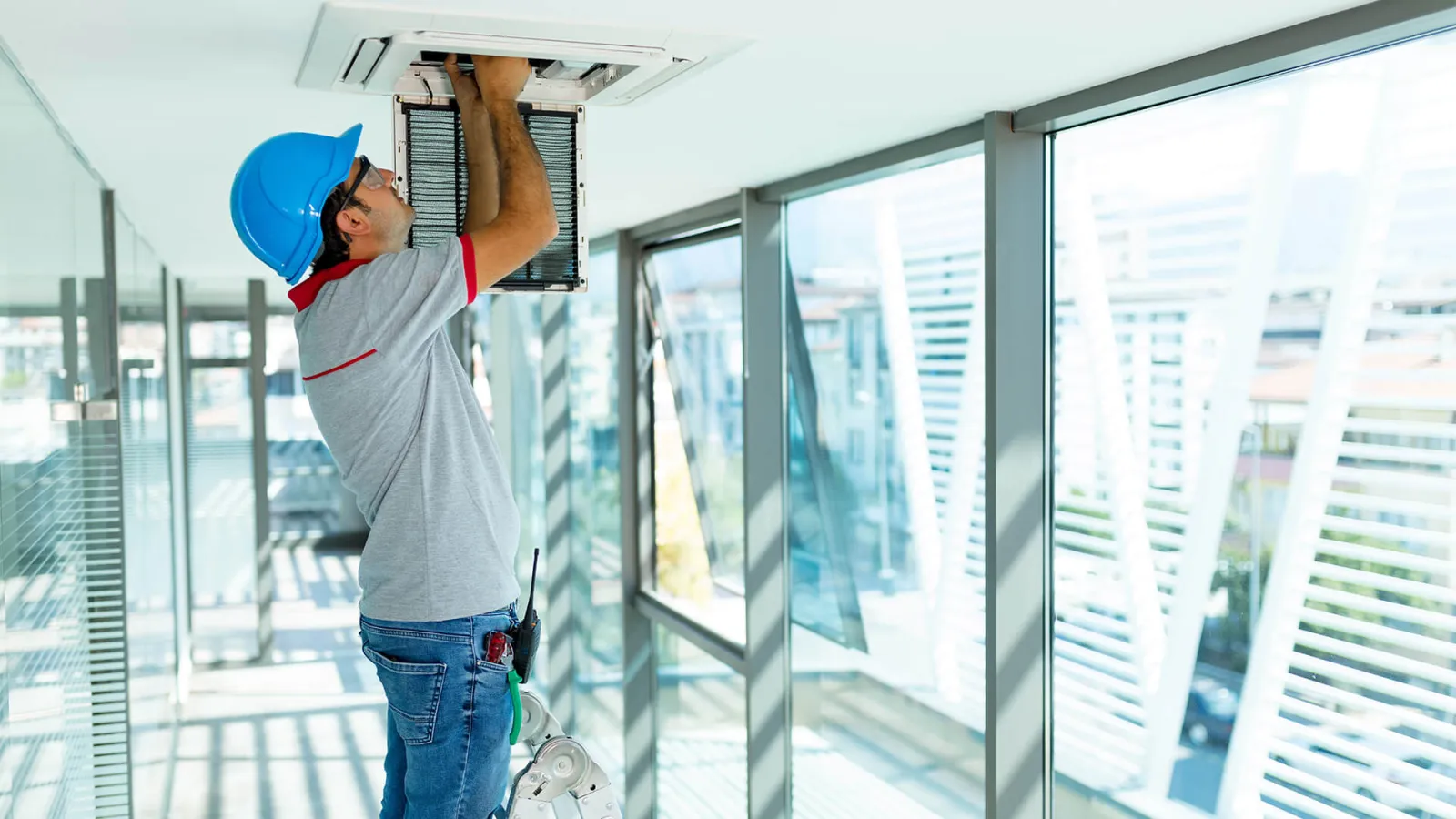 Technician in a blue hard hat repairing an air conditioning unit in a modern office with large glass windows.