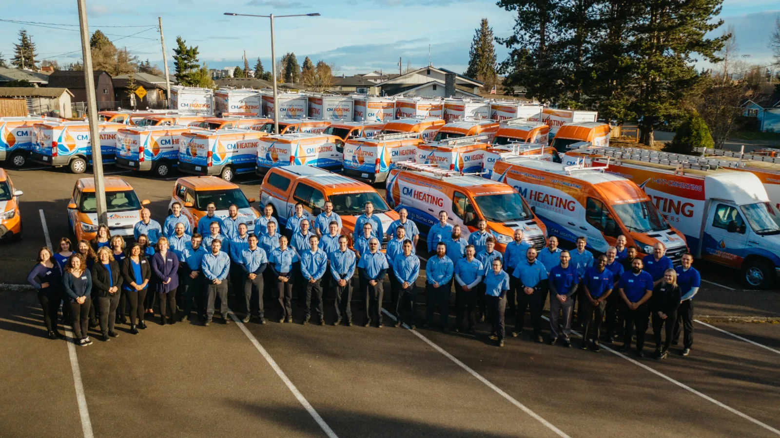 Large team of CM Heating employees standing in front of company branded vans in parking lot under blue sky