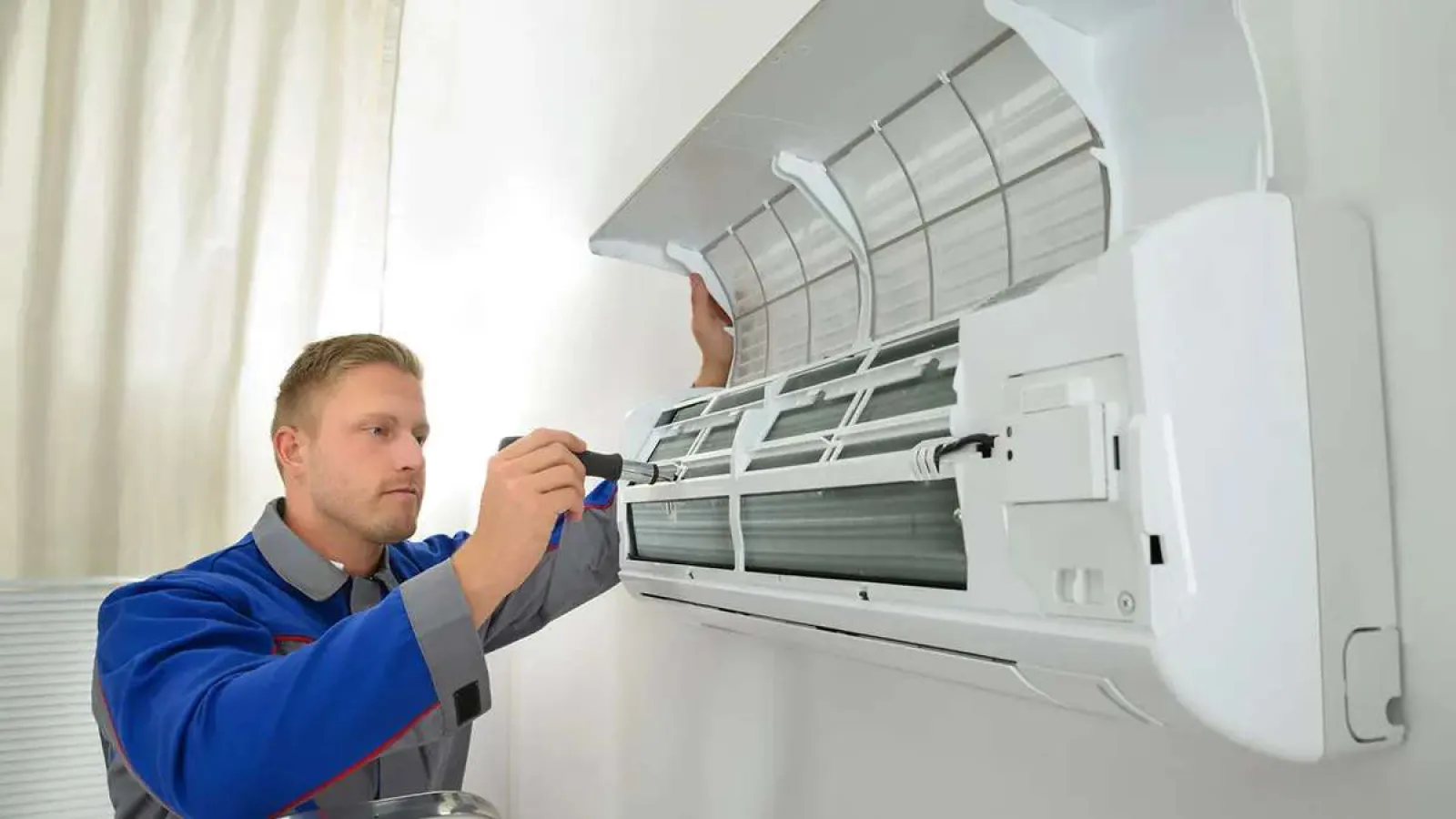 Technician repairing a wall-mounted air conditioner indoor unit with a screwdriver in a bright room.