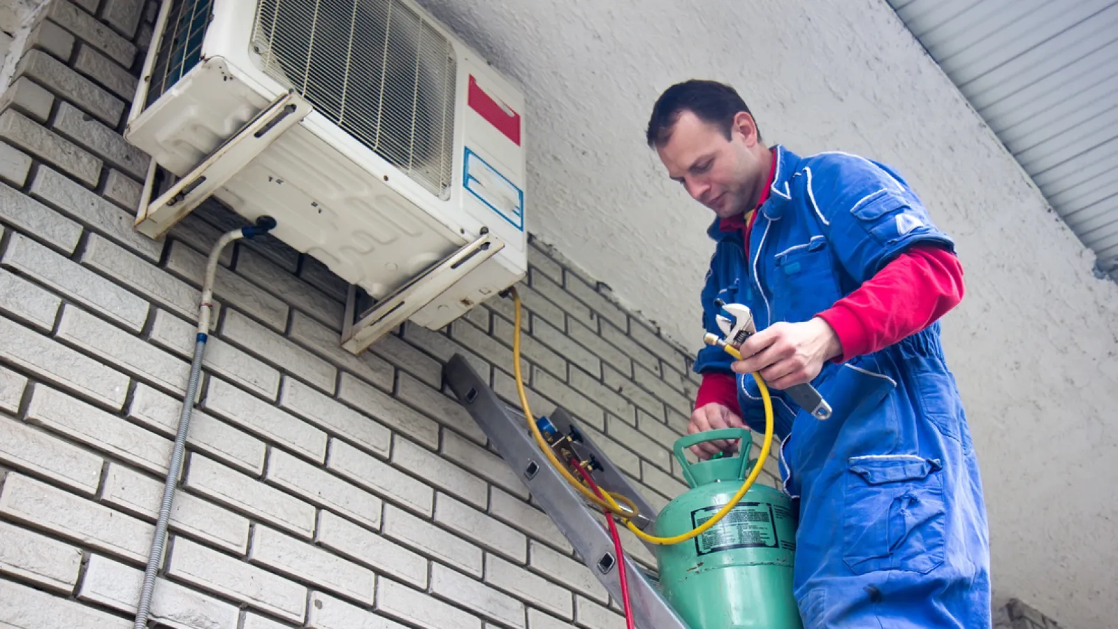 Technician in blue coveralls servicing an outdoor air conditioning unit with refrigerant tank on a ladder.