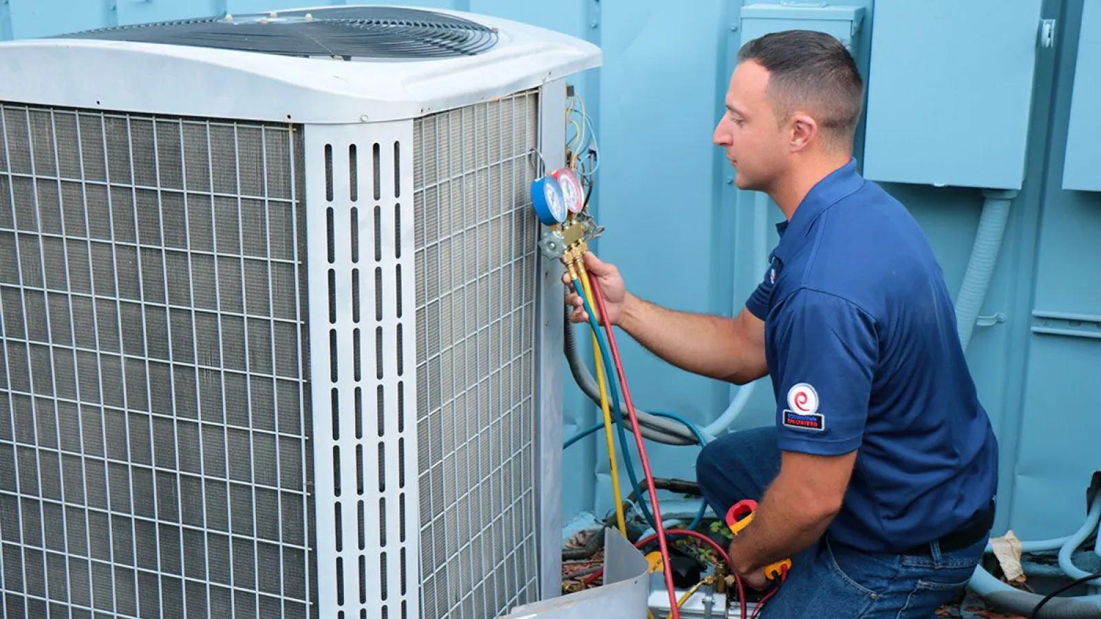 Technician in blue uniform repairing air conditioning unit using gauges and tools outdoors near blue wall.
