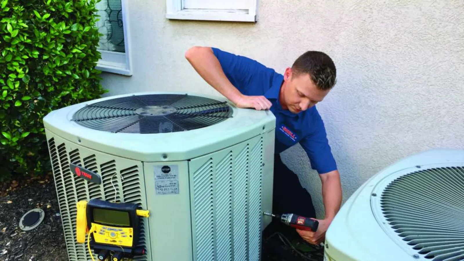 Technician repairing outdoor HVAC units with tools beside a house exterior wall and greenery.