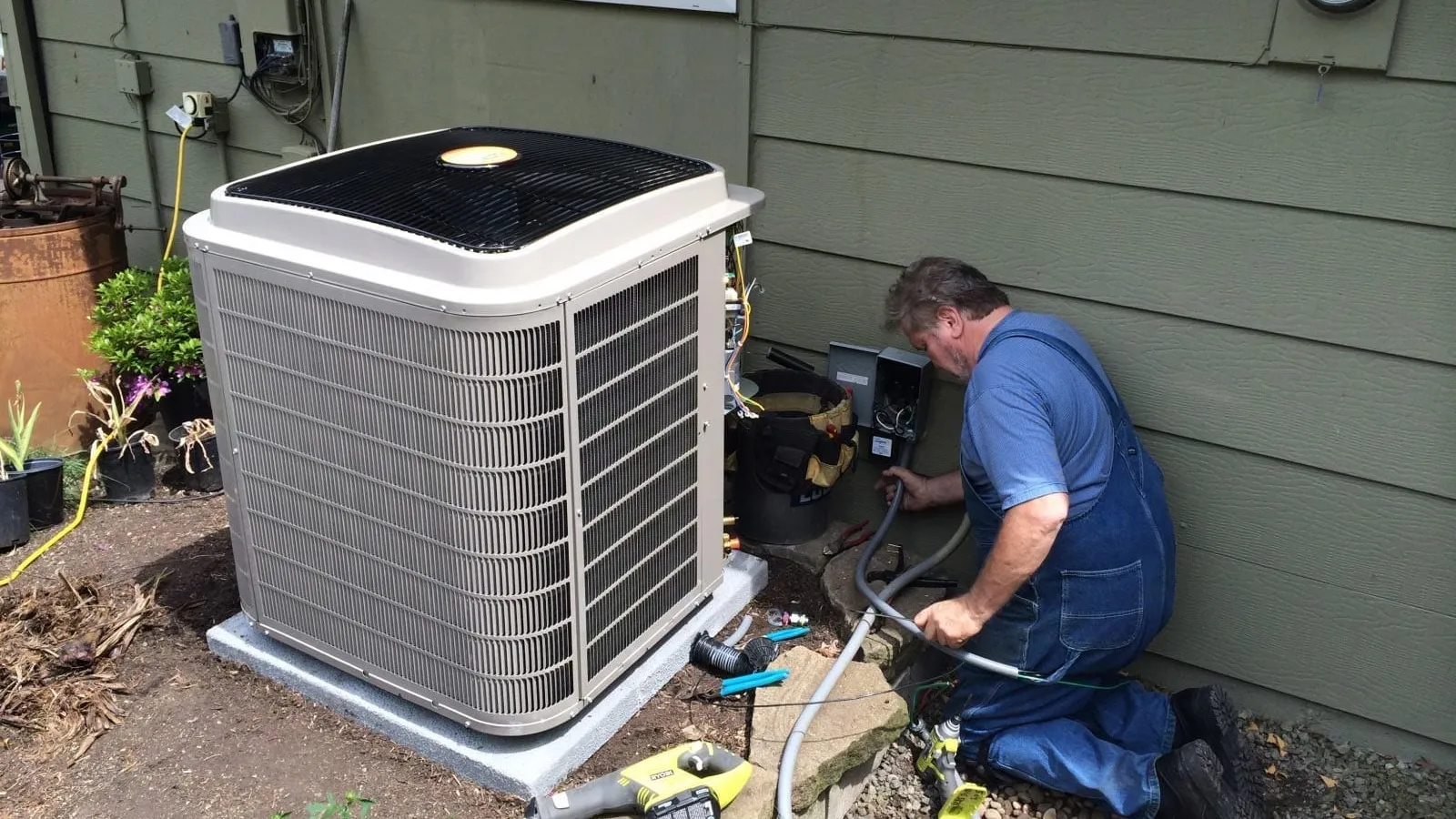 Technician in blue overalls repairing an outdoor air conditioning unit next to a house exterior wall.