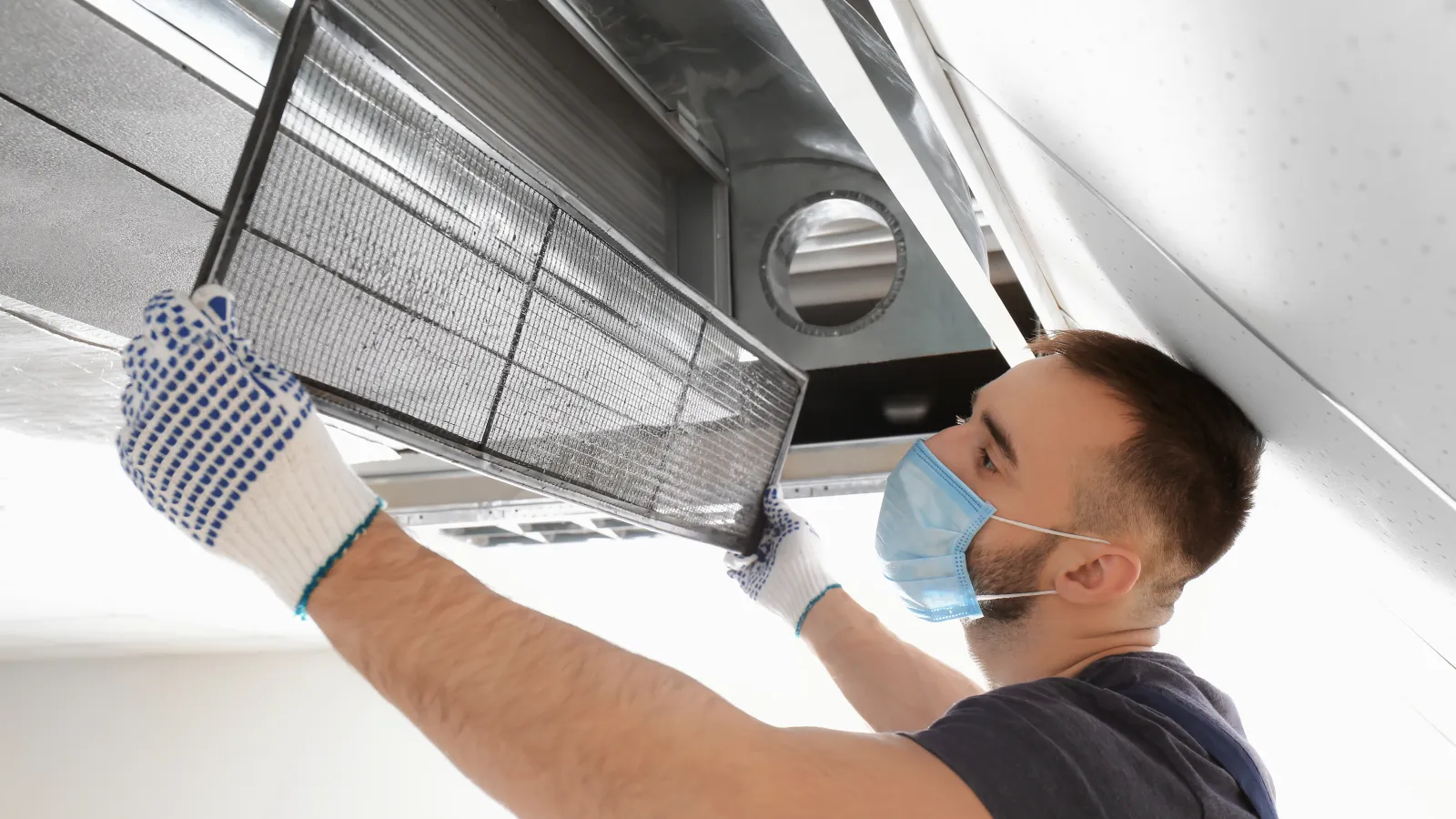 Technician wearing a mask and gloves replacing HVAC air filter in ceiling unit for maintenance