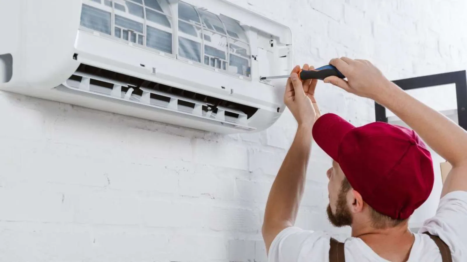 Technician in red cap using screwdriver to repair a wall-mounted split air conditioning unit indoors.