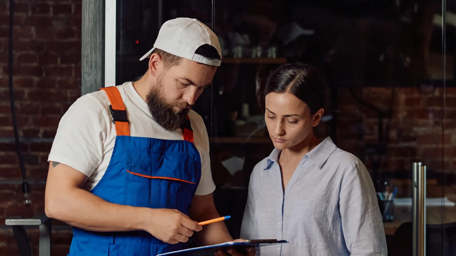Technician in blue overalls consulting with woman in casual clothes under a ceiling air conditioning unit.