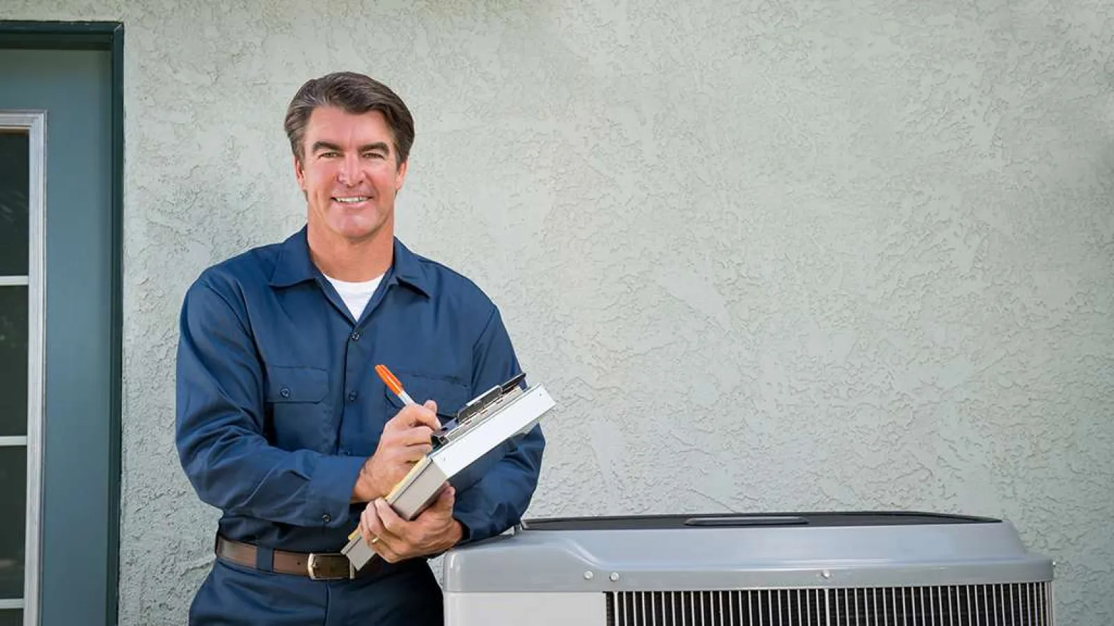 HVAC technician in blue uniform holding clipboard standing next to an outdoor air conditioning unit by a house wall.