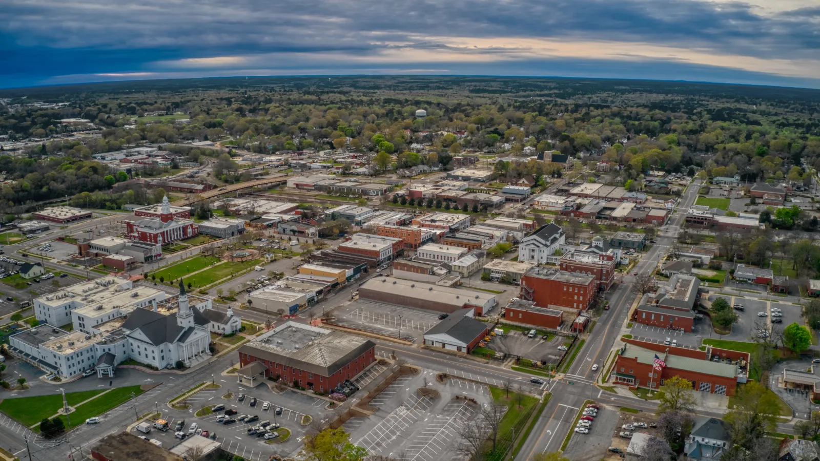 Aerial view of a small town with buildings, roads, parking lots, and surrounding greenery under a cloudy sky.