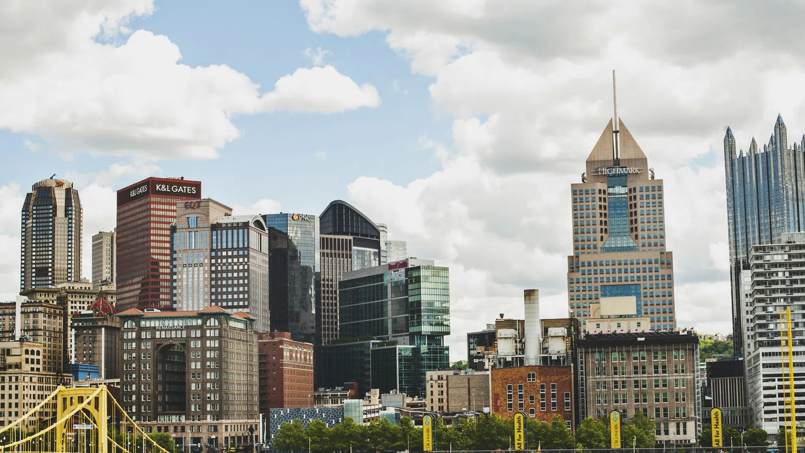 Pittsburgh city skyline with tall buildings, a yellow bridge, and partly cloudy sky during daytime.