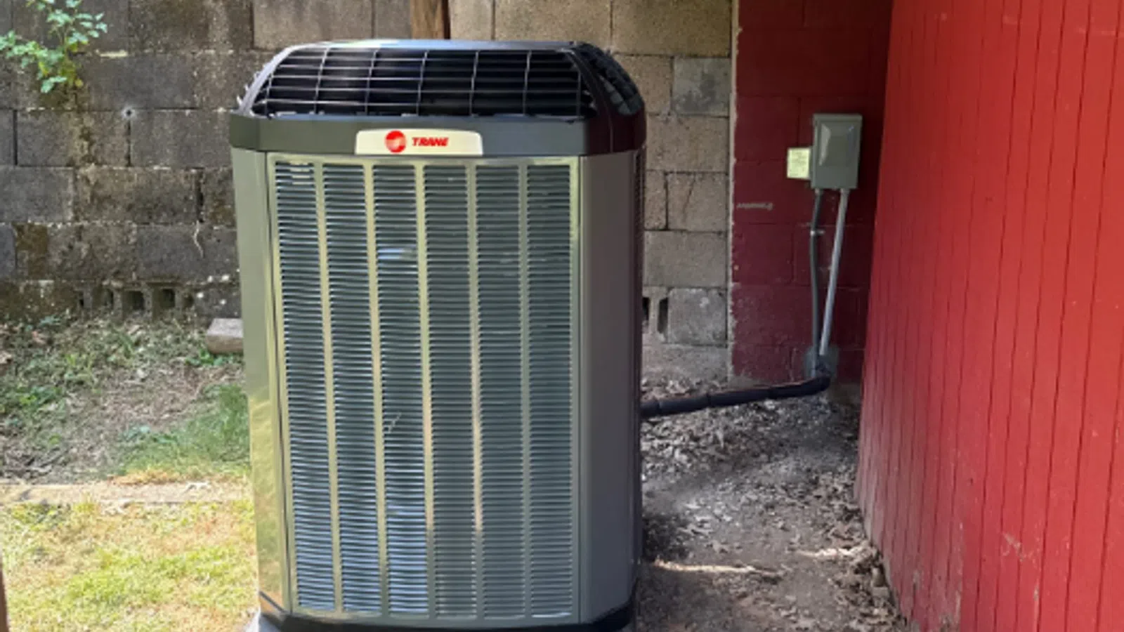 Outdoor Trane air conditioning unit installed beside a red wooden wall and concrete block fence on a concrete pad.