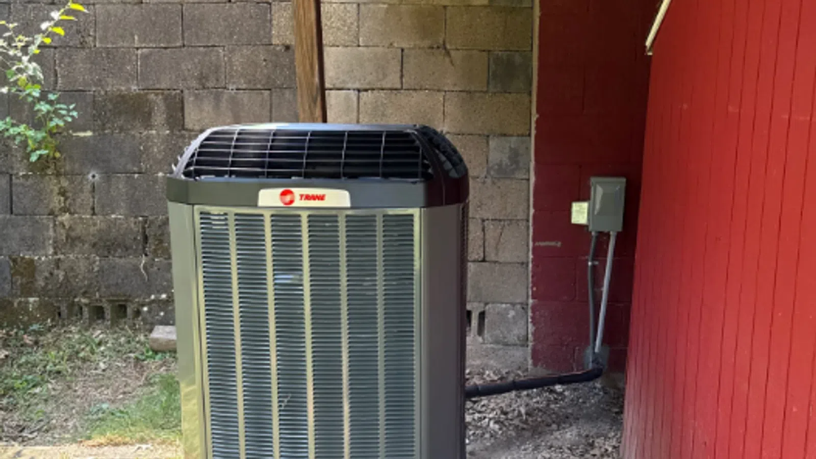Outdoor Trane air conditioning unit installed beside a red wooden wall and concrete block fence on a concrete pad.