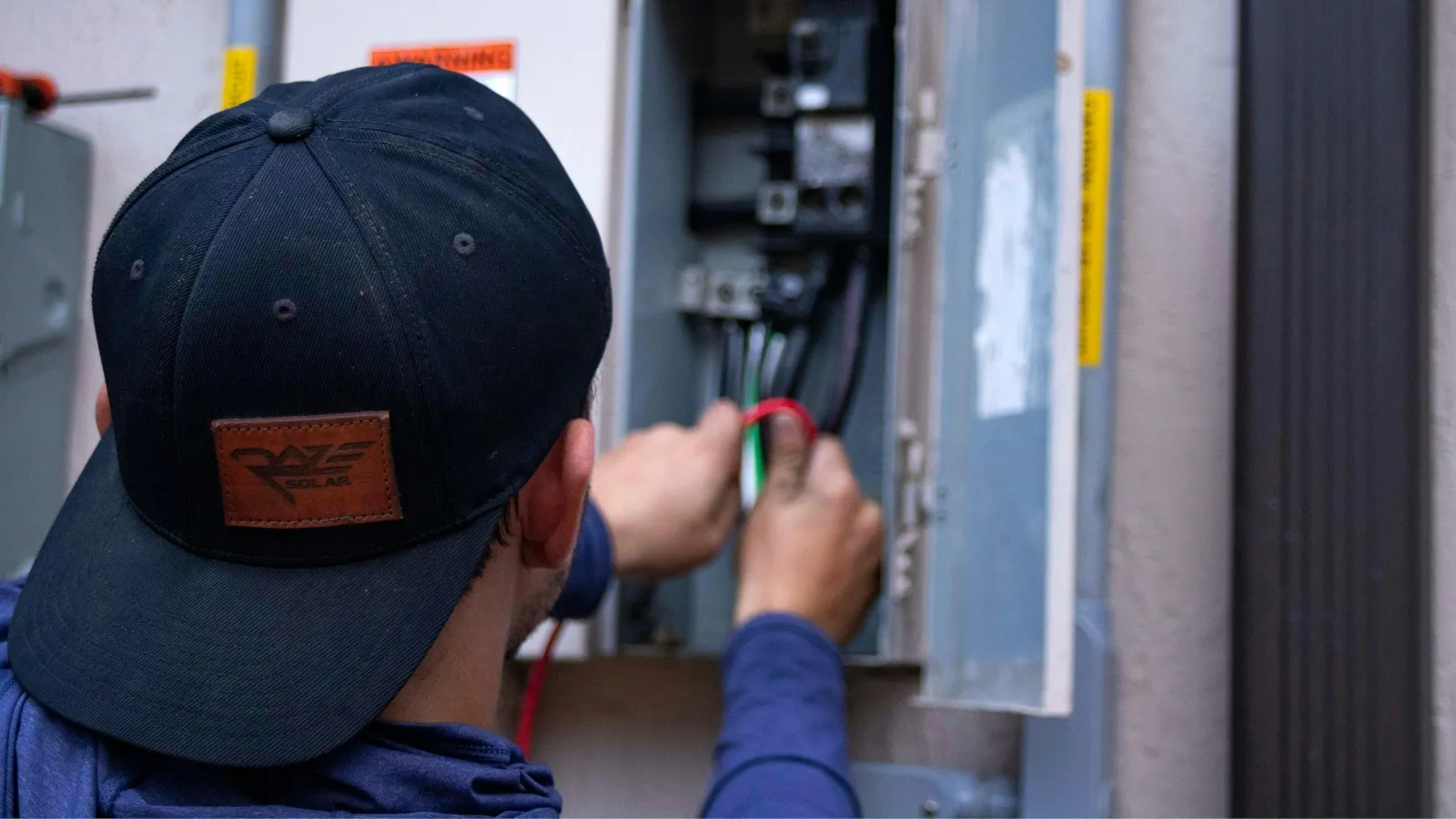 Electrician wearing a black cap fixing wiring inside an open electrical panel box outdoors