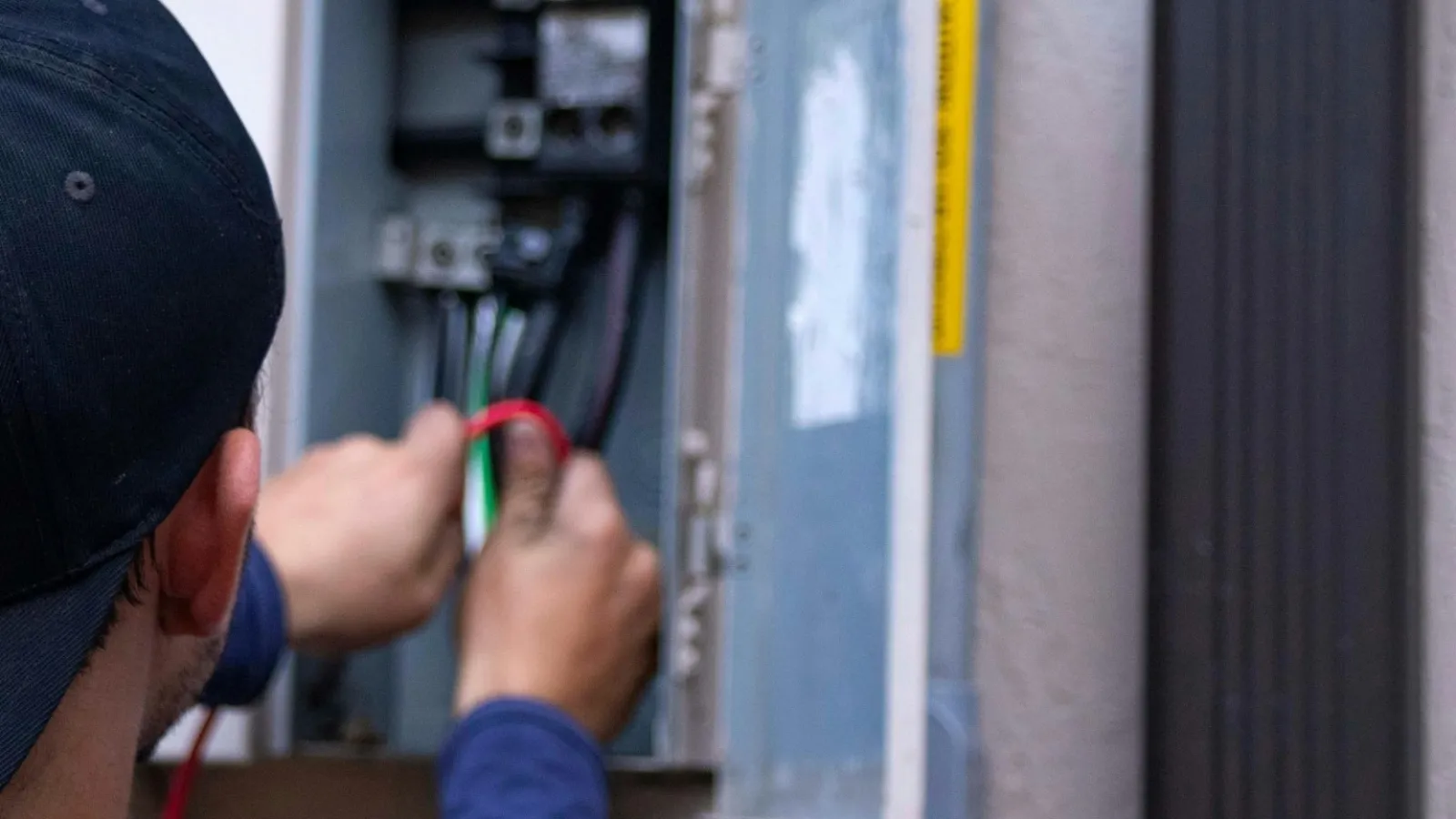 Electrician working on wiring inside an open electrical panel wearing a black cap and blue shirt