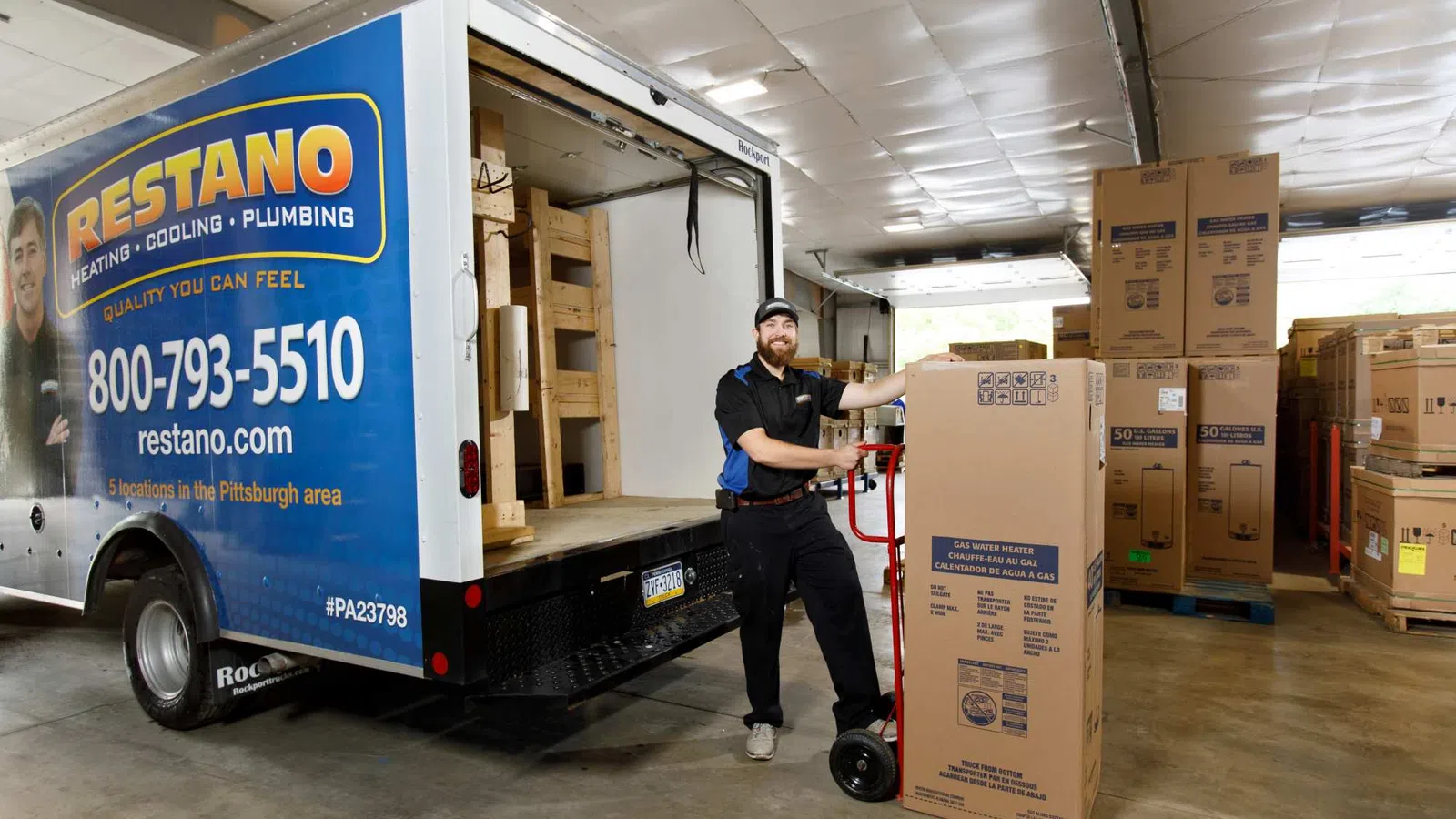 Delivery worker unloading large boxed gas water heater from Restano service truck inside warehouse.