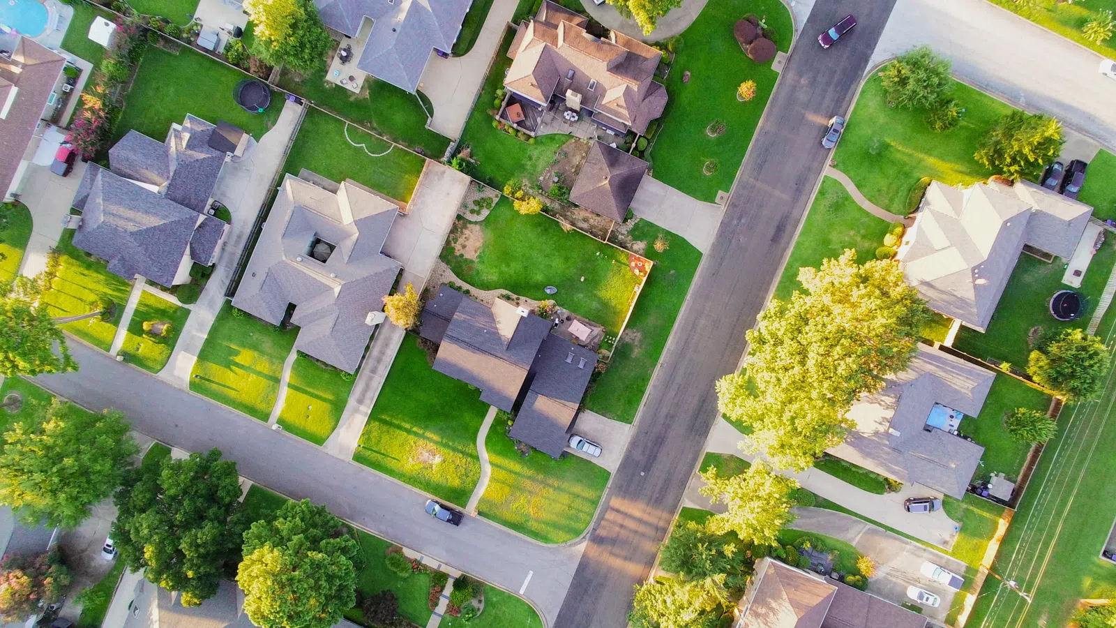 Aerial view of suburban neighborhood showing houses, green lawns, trees, and roads on a sunny day