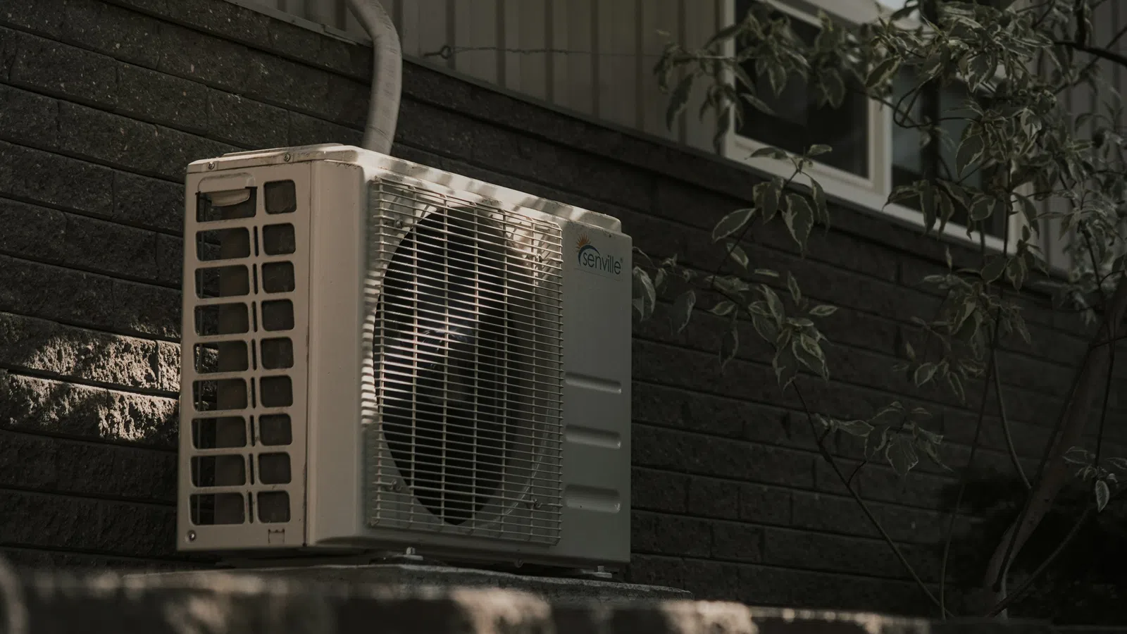 Outdoor HVAC unit mounted on a stone platform against a dark brick wall with foliage nearby.