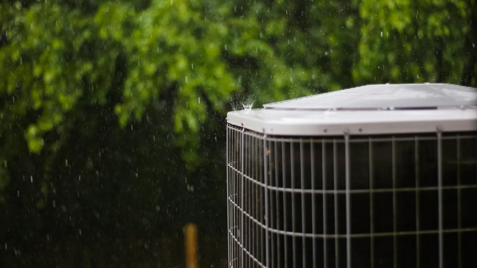 Outdoor air conditioning unit beside trees in the rain with water droplets falling on the metal surface.