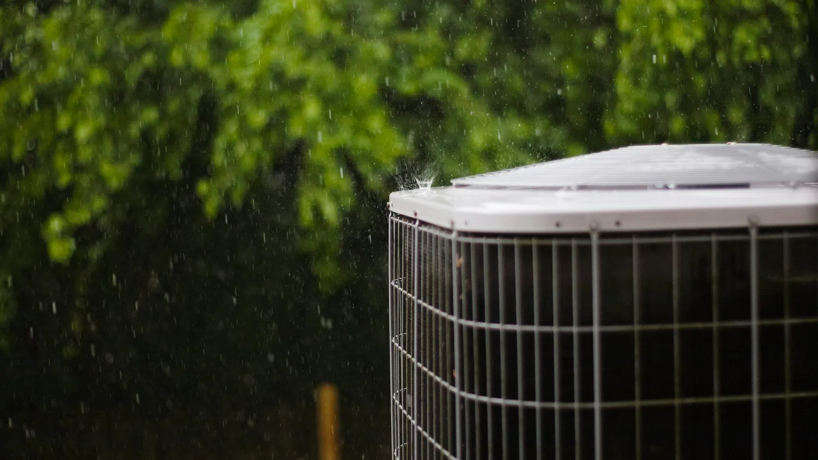 Outdoor air conditioning unit operating in rainy weather with green trees in the background.