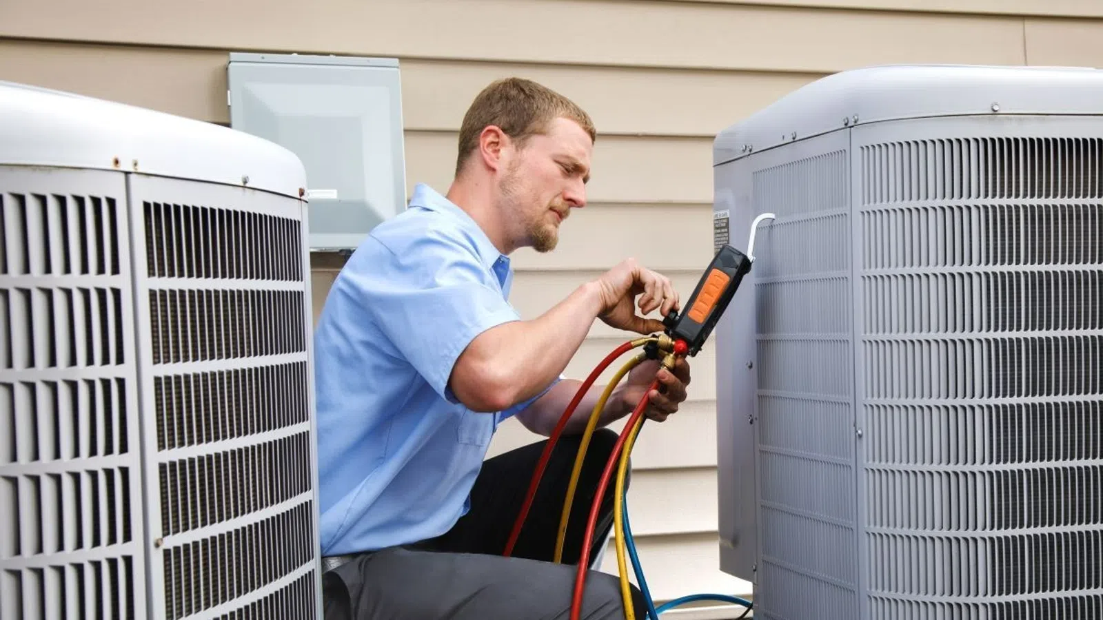 Technician in blue shirt using gauges to service HVAC unit outside a house.