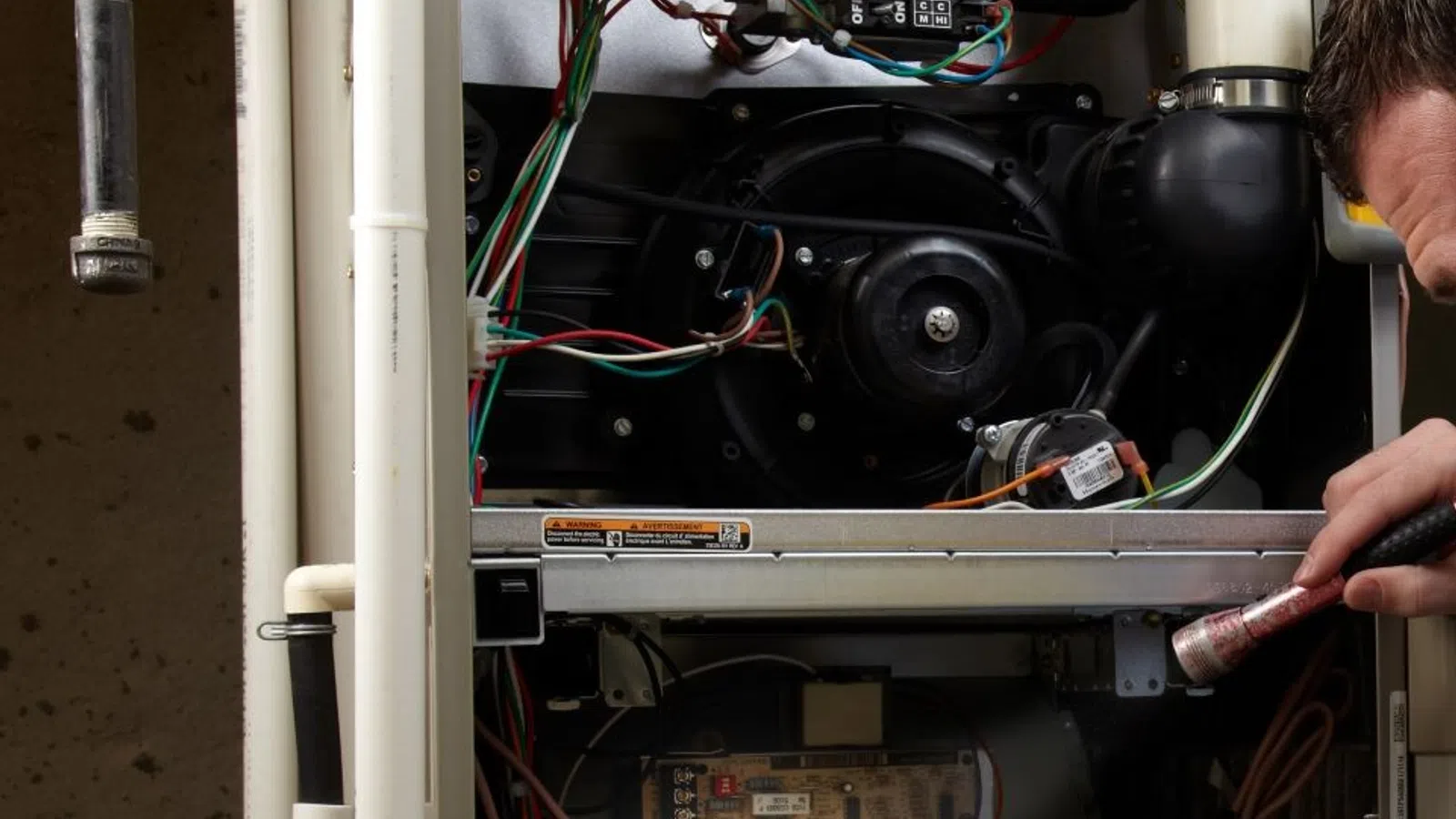 Technician servicing a furnace, inspecting electrical components with tools in a heating system.