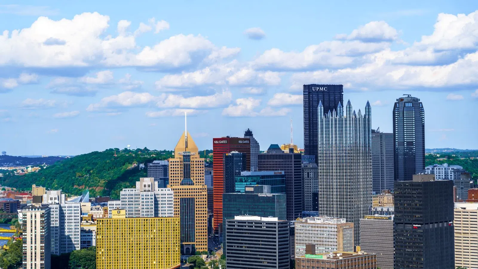 Skyline view of Pittsburgh with the UPMC building, green hills, and bright blue sky dotted with clouds.