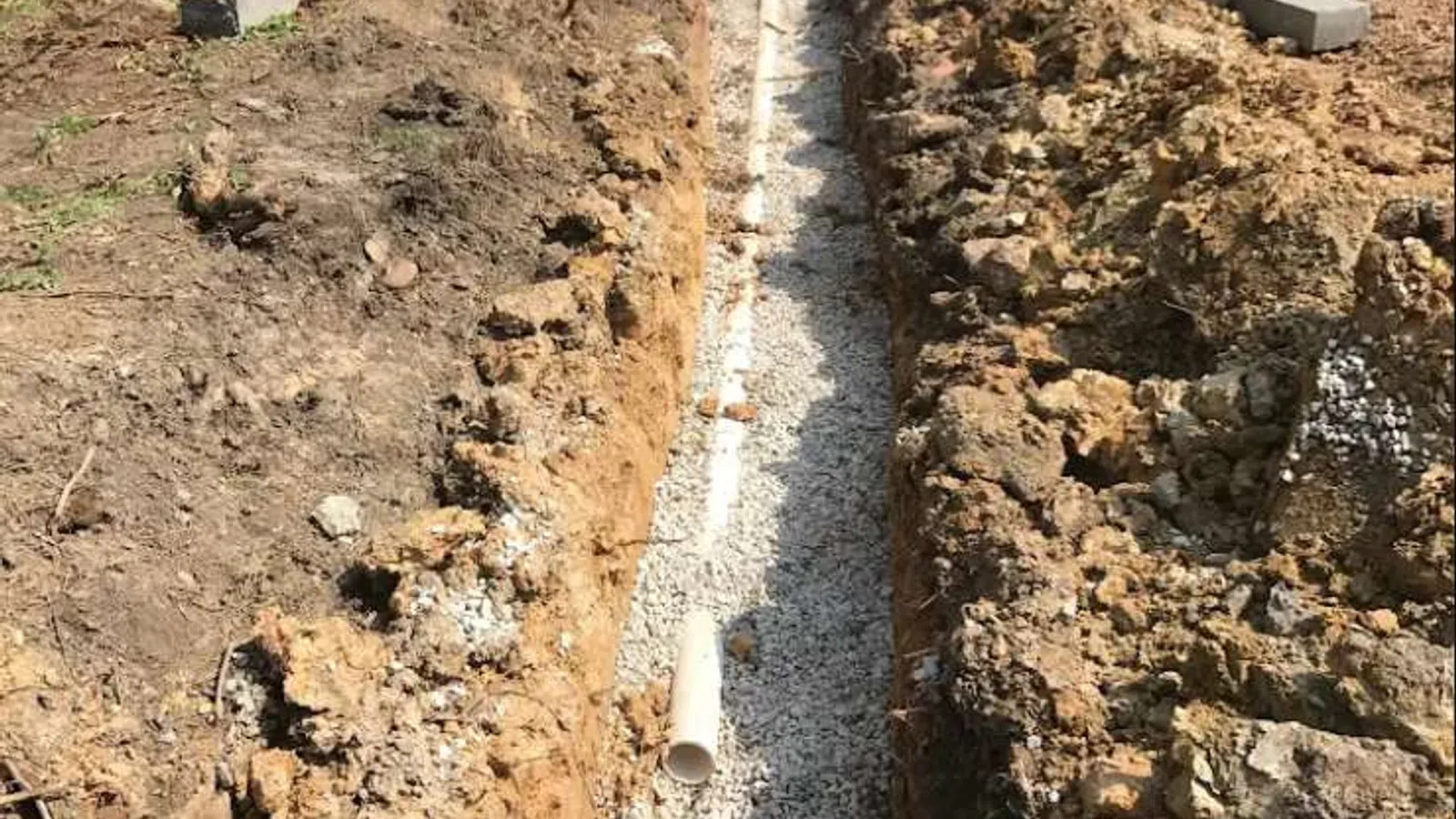 Construction site trench with gravel base and PVC pipe installation next to a brick building under excavation.