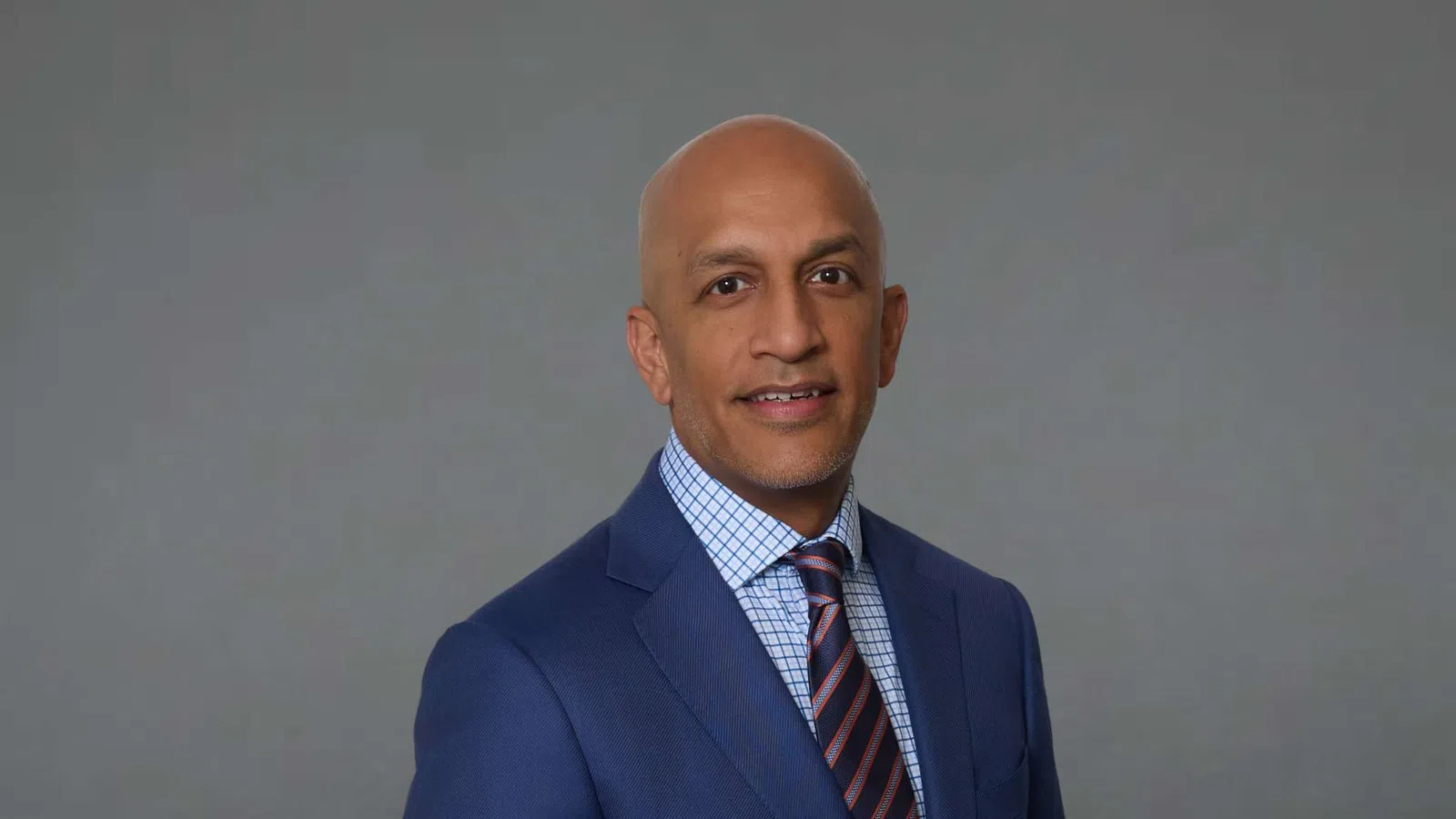 Professional man in a blue suit, checkered shirt, and striped tie standing against a gray background.