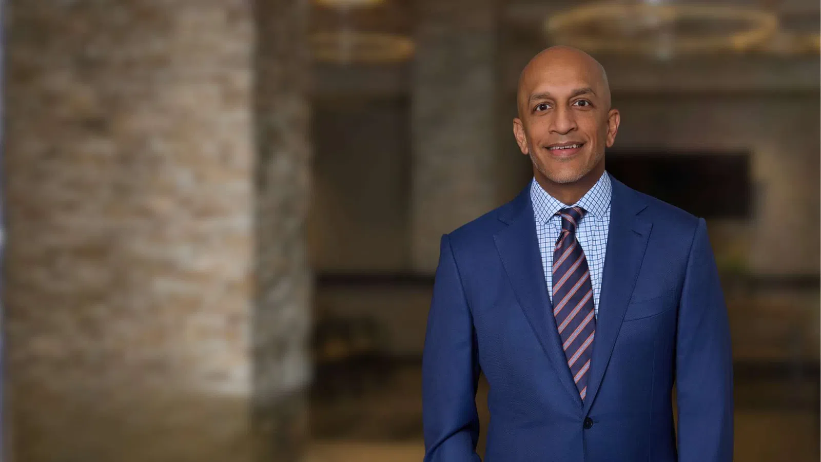 Professional man in blue suit and striped tie smiling in a blurred indoor setting with stone walls.
