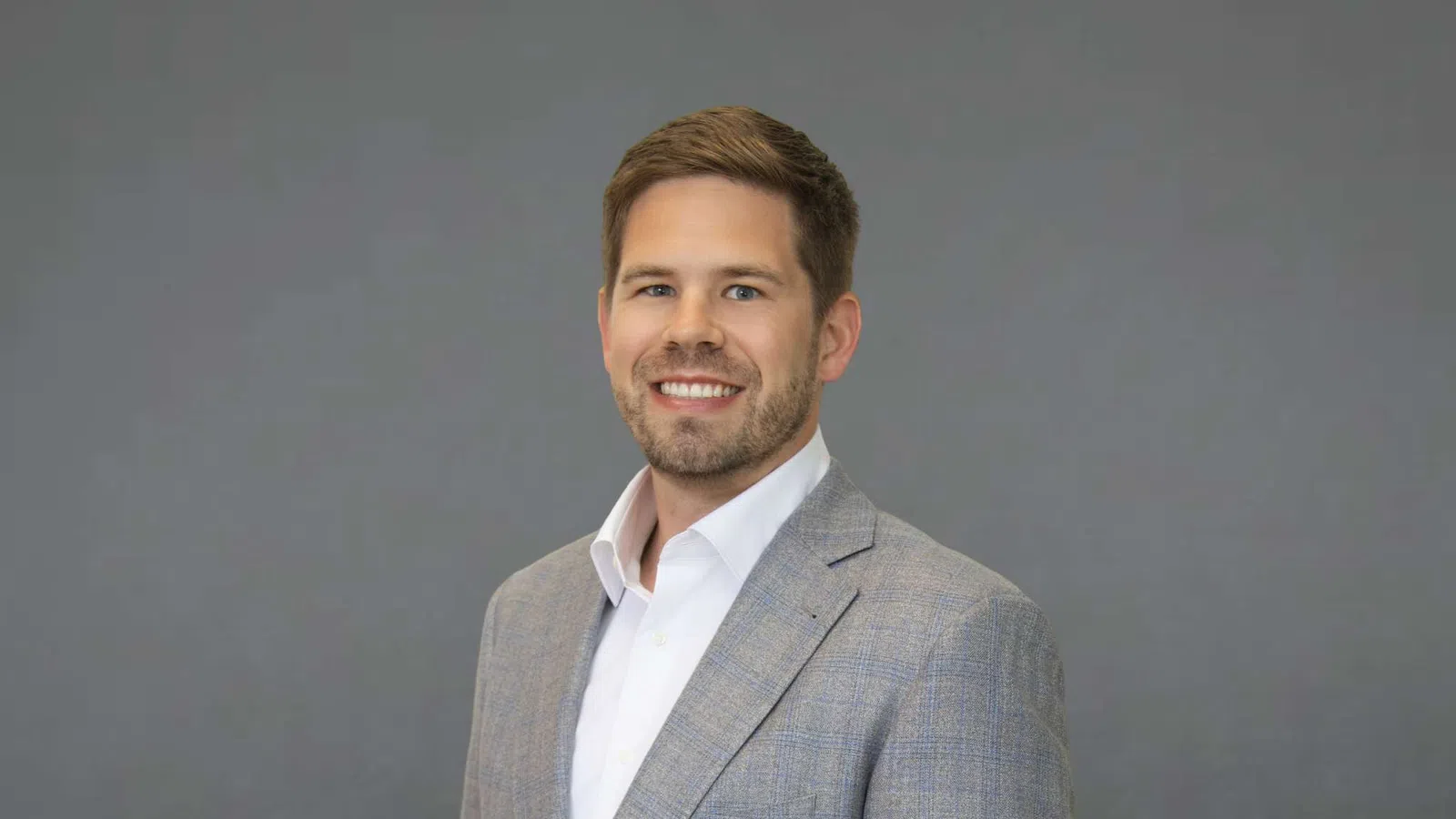 Smiling man in gray suit jacket and white shirt against a plain gray background posing for portrait.