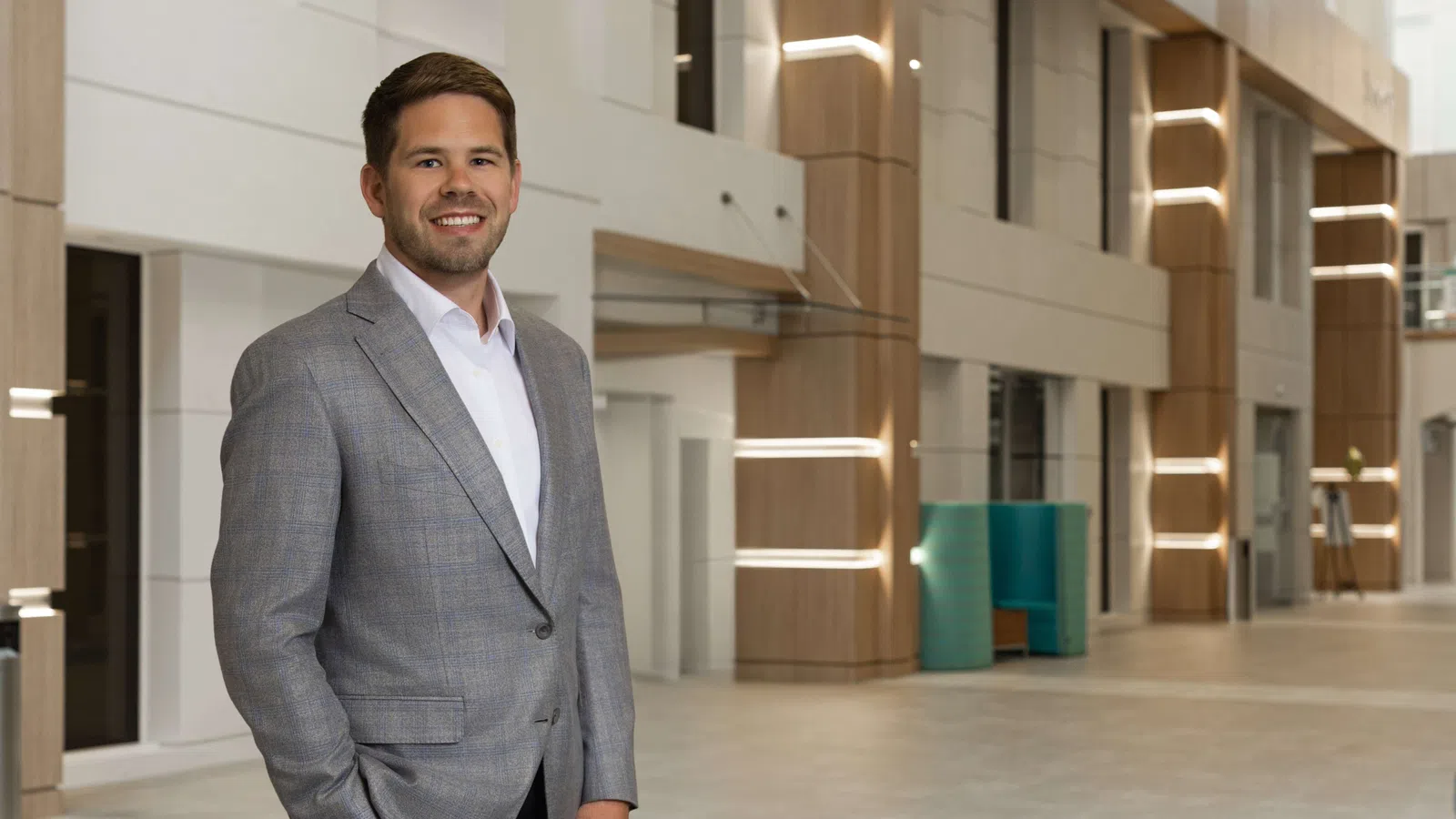 Smiling businessman in gray blazer standing in modern office lobby with wooden accents and soft lighting.