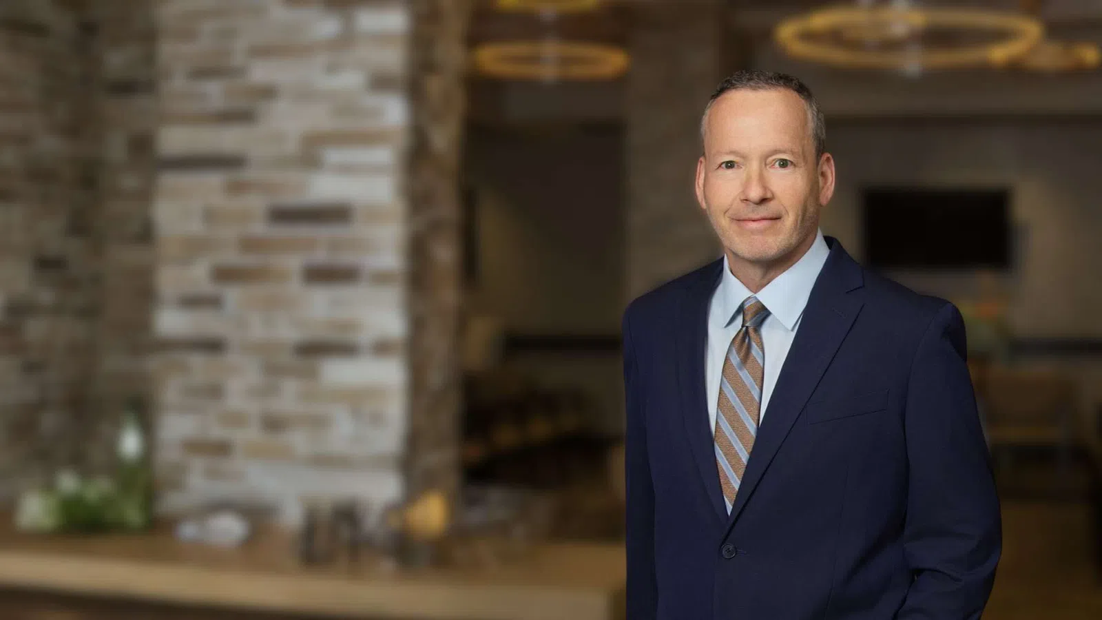 Professional man in blue suit and striped tie standing in modern office with brick wall background.