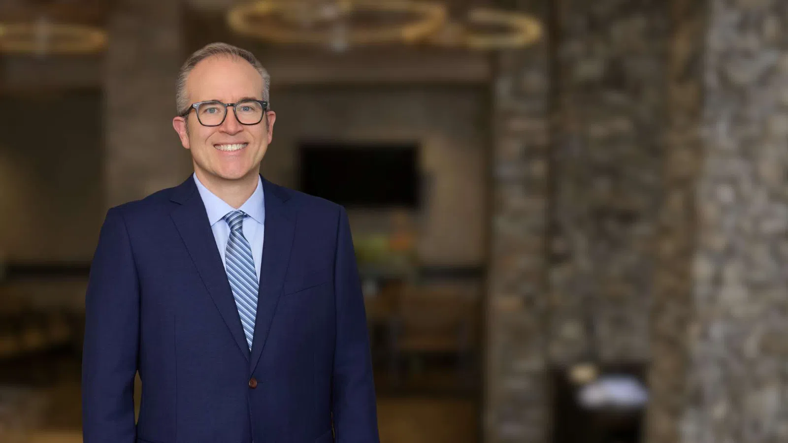 Smiling man in blue suit and glasses standing indoors with stone wall and chandelier background.