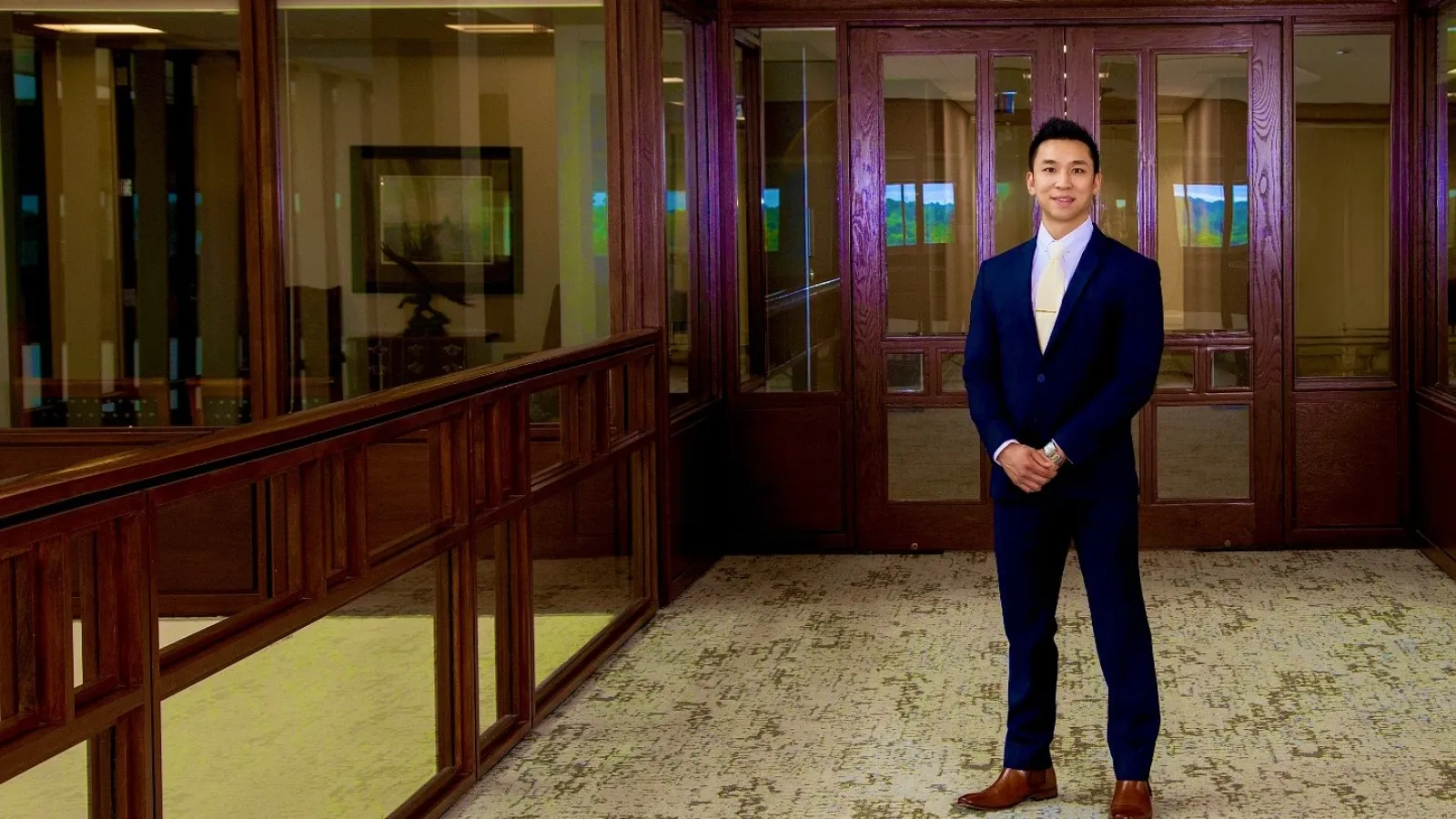 Man in blue suit standing in a spacious corridor with wood-paneled walls and glass doors.