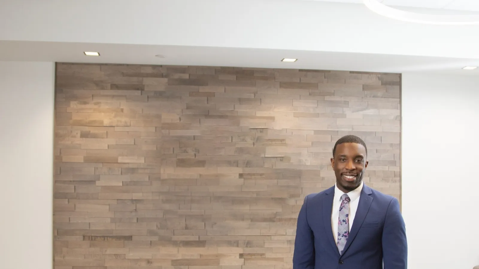 Professional man in a blue suit standing and smiling in a modern office lounge with wood accent wall.