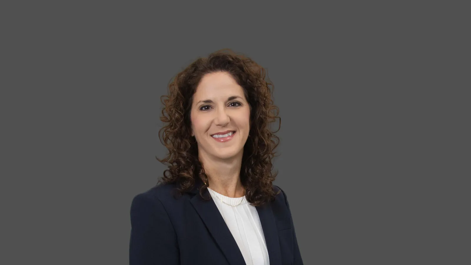 Professional woman with curly hair wearing a dark blazer and white blouse against a gray background.