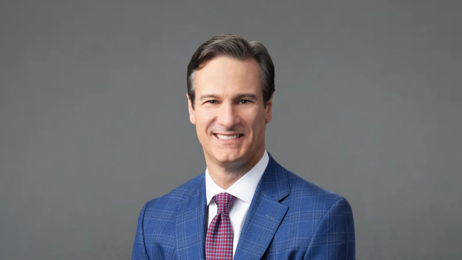 Man in blue checkered suit with red tie smiling against gray background studio portrait.