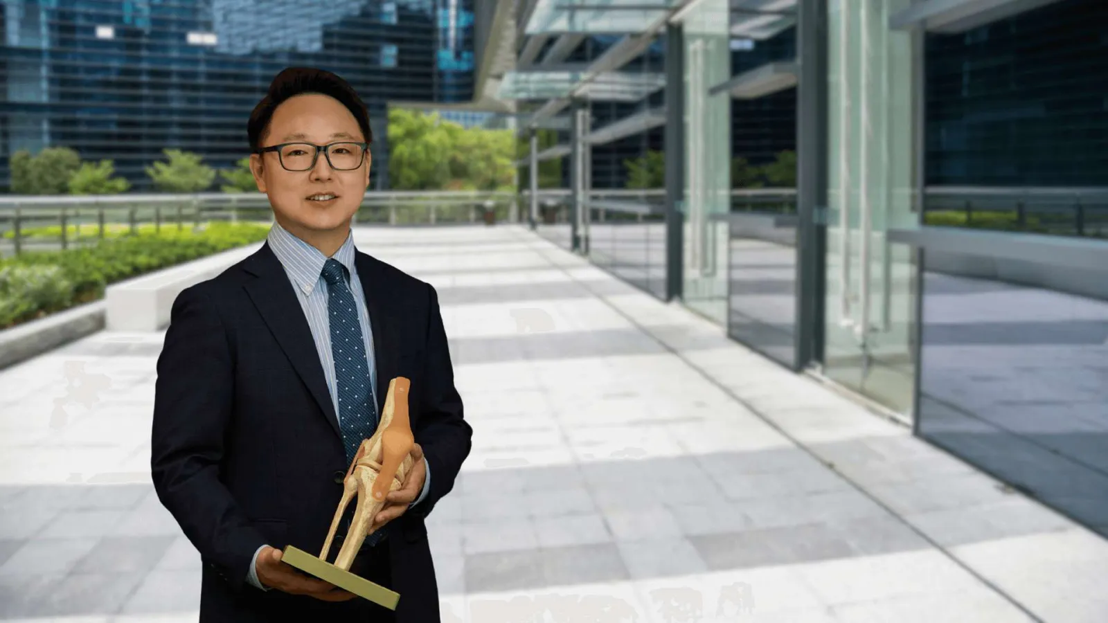 Man in suit holding anatomical knee joint model outdoors near modern office buildings under sunlight.